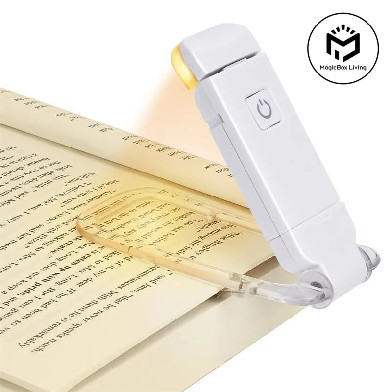 A white handheld book light with a clear clip is shown resting on an open book, illuminating the text with a warm yellow glow. The MagicBox Living logo is visible in the top right corner.